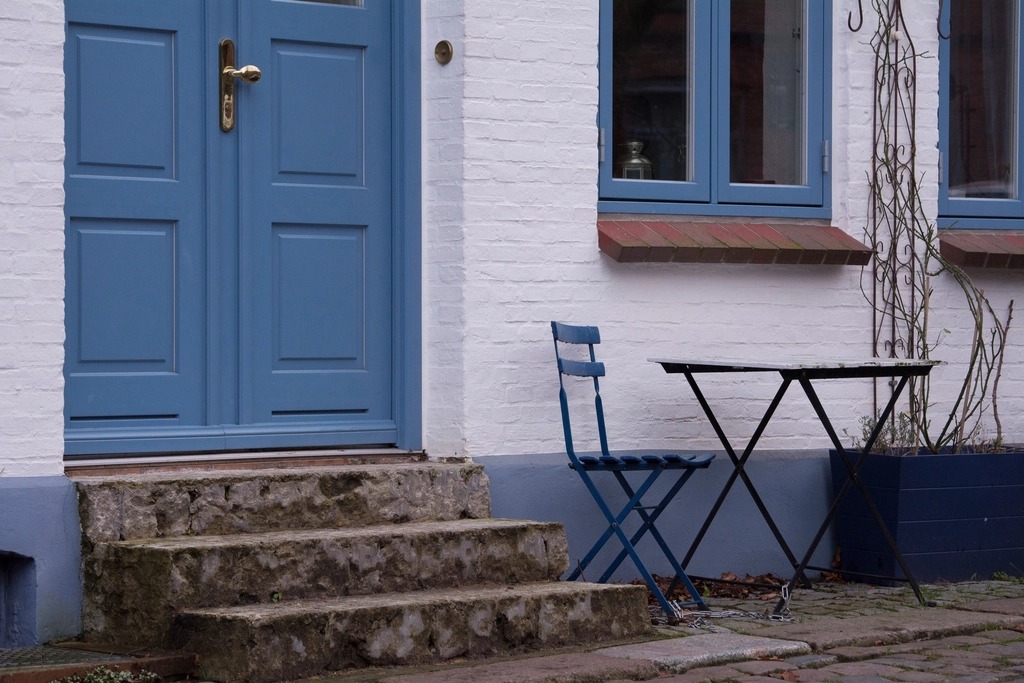 Blue-gray paneled front door above stone steps beside a white brick wall and a blue bistro chair