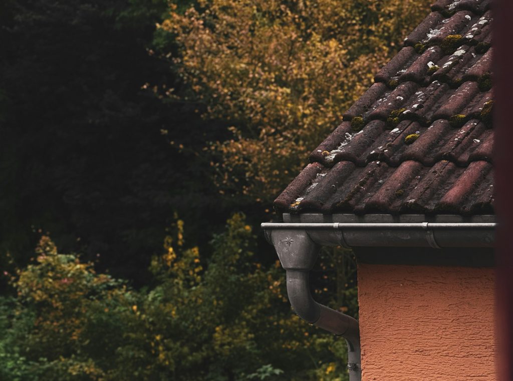 Metal gutter and downspout along a roof edge with autumn trees turning yellow and orange in the background