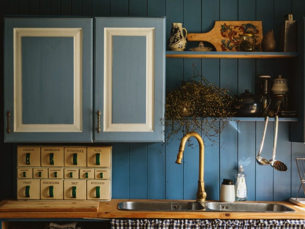 Smoky blue painted kitchen cabinets with cream door panels above a butcher-block counter, brass faucet and blue beadboard walls