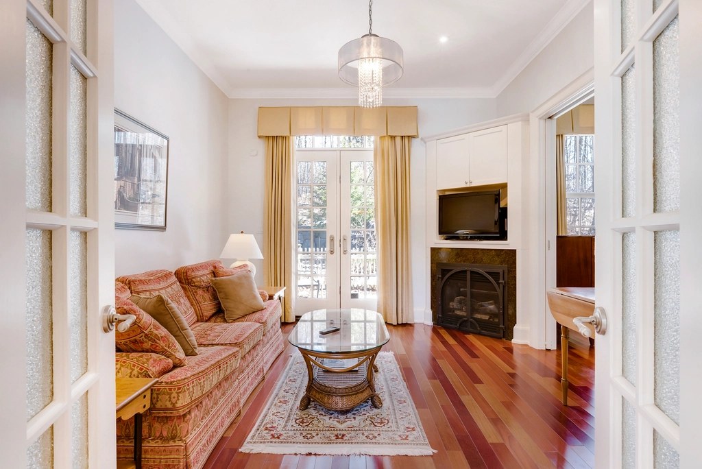 Sitting room with warm white walls, white trim and built-ins, a rust-patterned sofa, cherry wood floors and a fireplace beside French doors