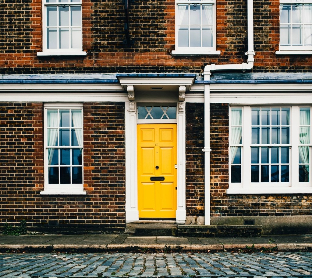 Bright yellow paneled front door with white trim and a transom window on a dark brick house beside a cobblestone street